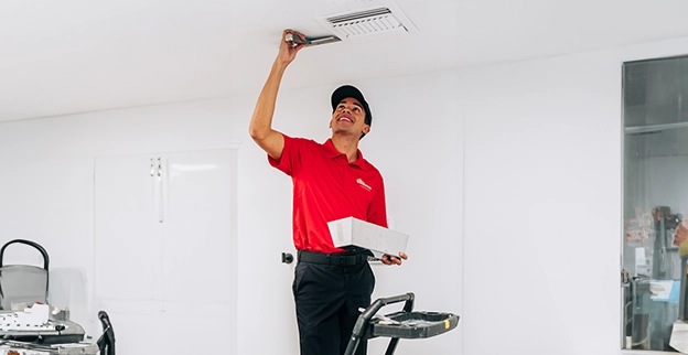 A worker in a red shirt uses a tool to adjust a ceiling vent in a clean, empty room.