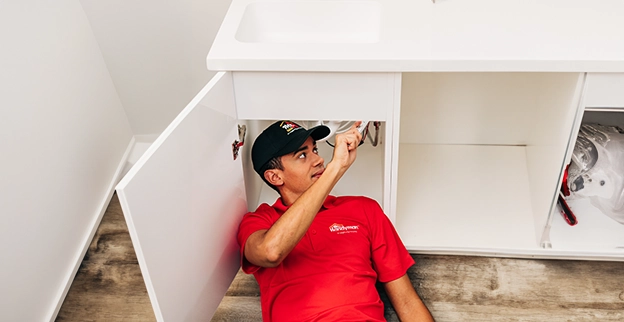 A technician in a red shirt works under a kitchen sink.