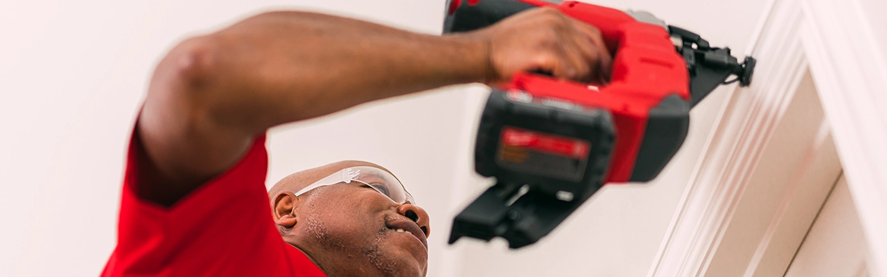 A man wearing glasses uses a power nail gun to attach trim to a wall focused on his task in a bright indoor setting.