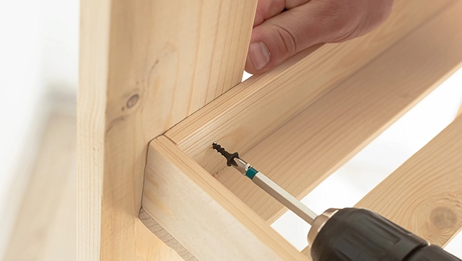 A person using a power drill to screw wooden planks together focusing on the join between two pieces of light-colored wood.