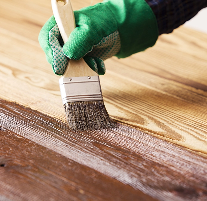 A gloved hand applies wood finish with a brush on a wooden surface.