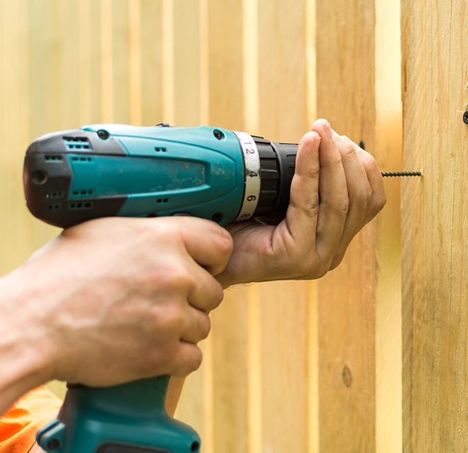A person using a cordless drill to insert a screw into a wooden fence.