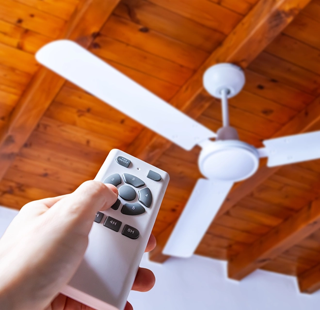 A hand using a remote control to adjust a ceiling fan against a wooden ceiling backdrop.