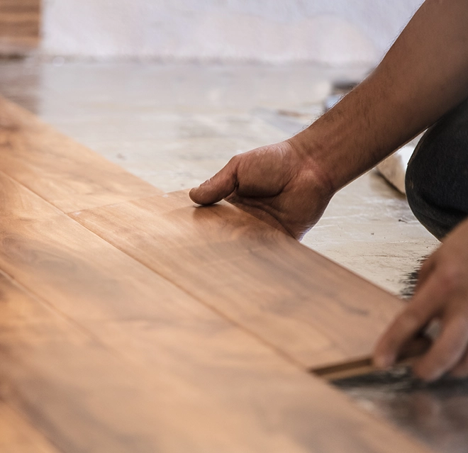 A close-up of a person's hands installing wooden flooring planks on a smooth surface.