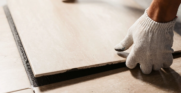 A person wearing a white glove carefully places a large tile onto a freshly laid floor.