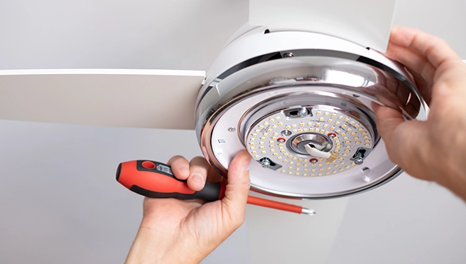 A person holds a screwdriver while installing a light fixture on a ceiling fan.