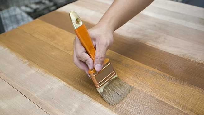 A hand holding a brush applies a warm wood finish to a smooth wooden surface.