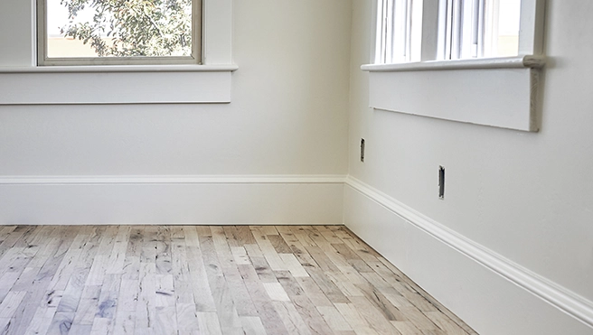 Bright, empty room with light wood flooring, white walls, and windows letting in natural light.