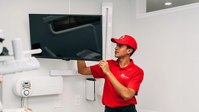 A technician in a red uniform installs a flat-screen TV on a white wall in a contemporary room.